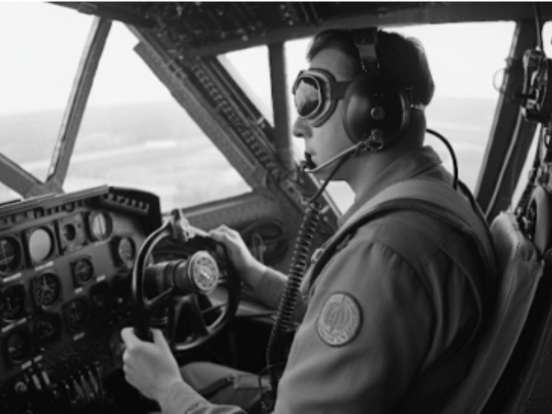 Black and white photograph of a pilot wearing a helmet goggles and headset operating the controls of an aircraft in flight