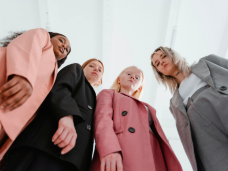 Four women in business attire stand close together, looking down at the camera. They wear various shades of suits, ranging from light pink to gray and black