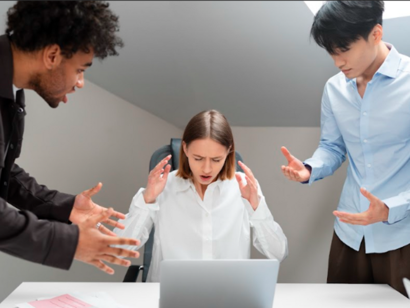 A person at a laptop with their back to the viewer and hands near their head, surrounded by workmates gesturing in frustration, depicting workplace stress.