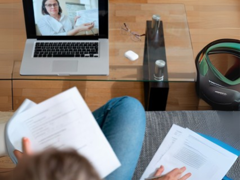 A person sits on a couch, looking at a laptop screen displaying a therapist during an online therapy session. The person holds papers, suggesting discussion or note-taking.